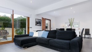 A modern living room with a sectional sofa, a dining area with a table and chairs, and a large window with a view of greenery. - YourHouse Cardinal (Can Picafort)