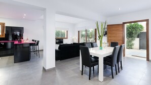 A modern kitchen with a black island, a dining area with a white table and black chairs, and a black sofa in the living area. - YourHouse Cardinal (Can Picafort)