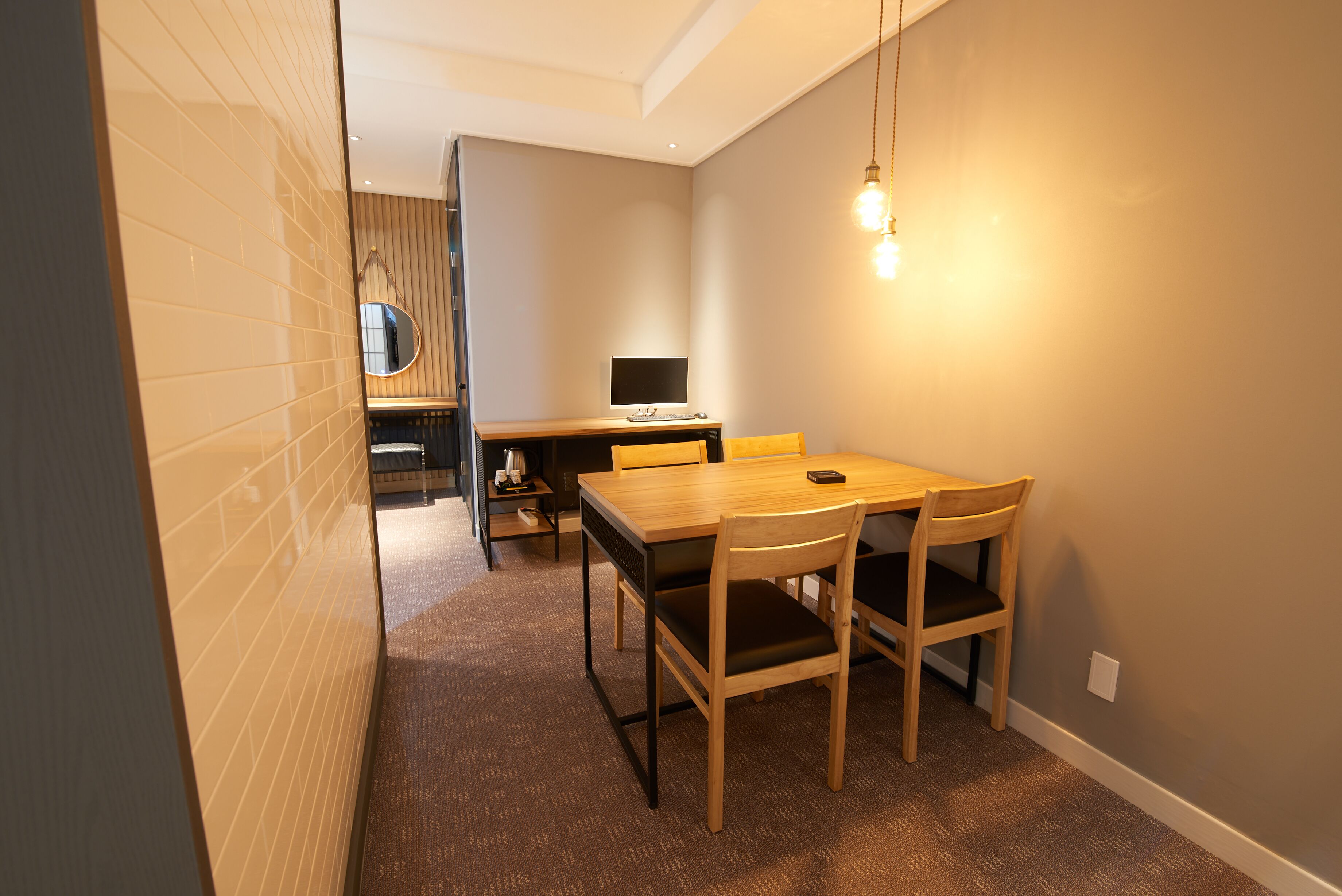 A compact room with a wooden dining table and chairs, a small desk with a computer, and a hanging light fixture.
