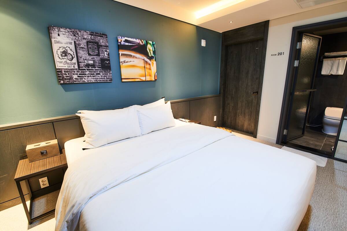 Una habitación de hotel con una cama grande, una mesita de noche con una caja encima, dos cuadros en la pared y una puerta al baño.