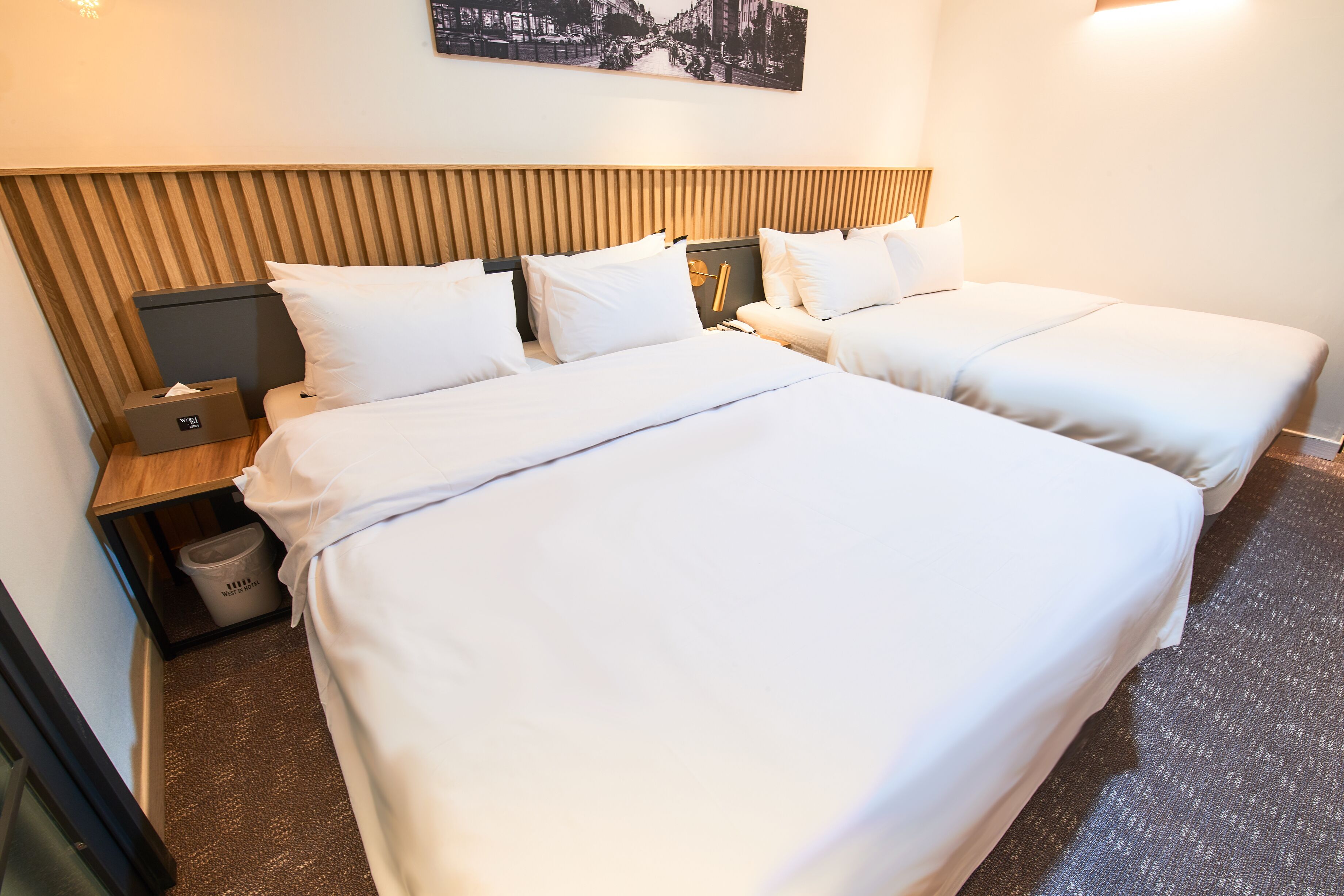 A hotel room with two beds, a wooden headboard, a waste bin, and a framed picture on the wall.