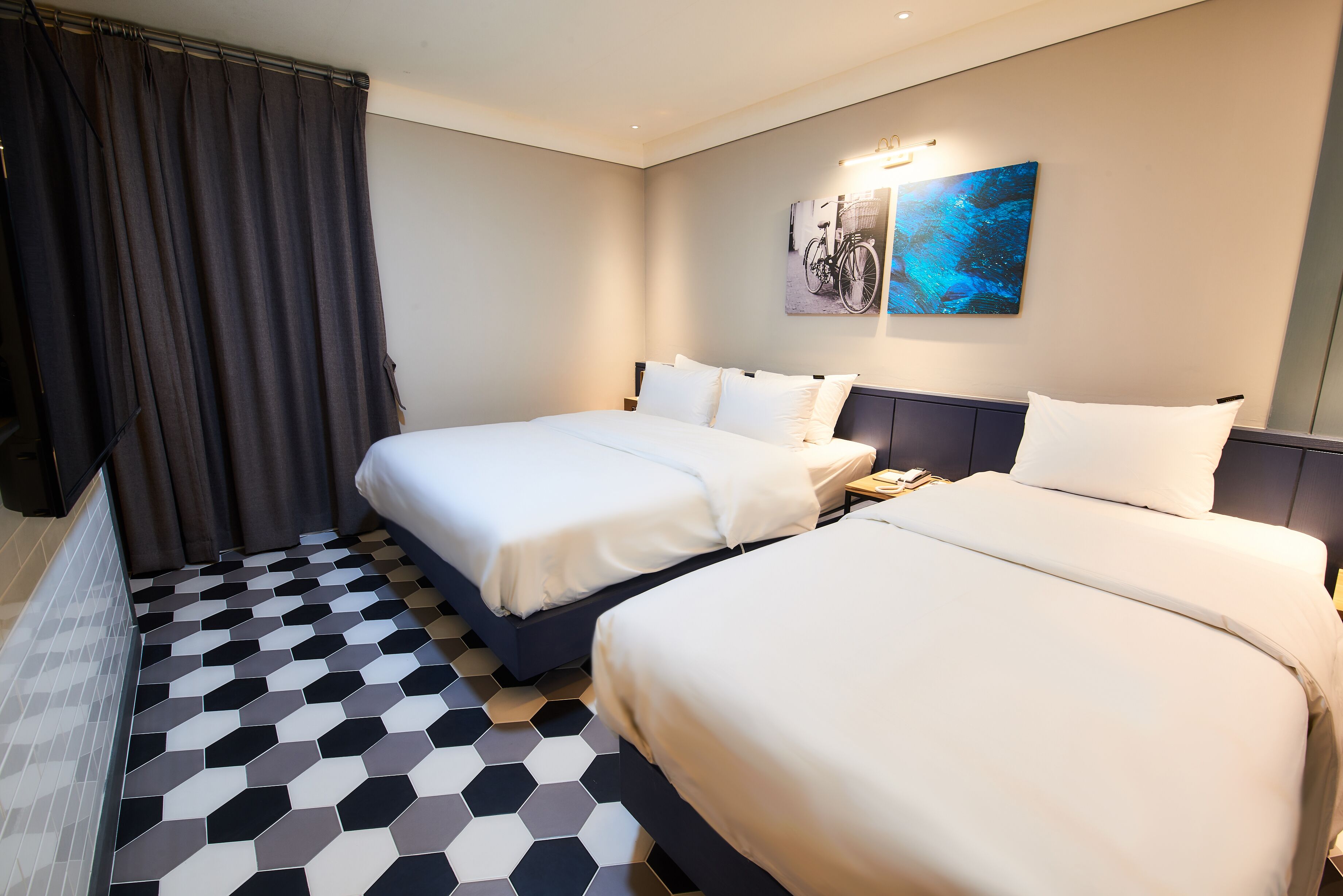 A hotel room with two beds, a large window with curtains, a wall-mounted artwork, and a geometric patterned floor.