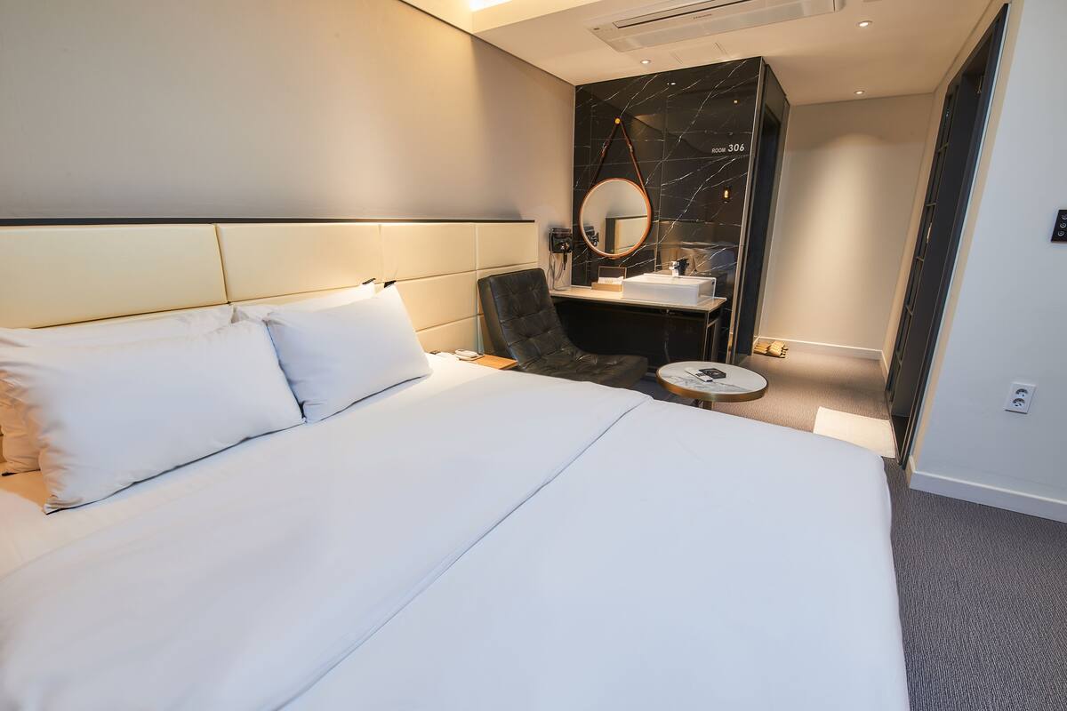 Una habitación de hotel con una cama grande, un sillón de cuero negro, un espejo redondo y una pequeña mesa redonda.