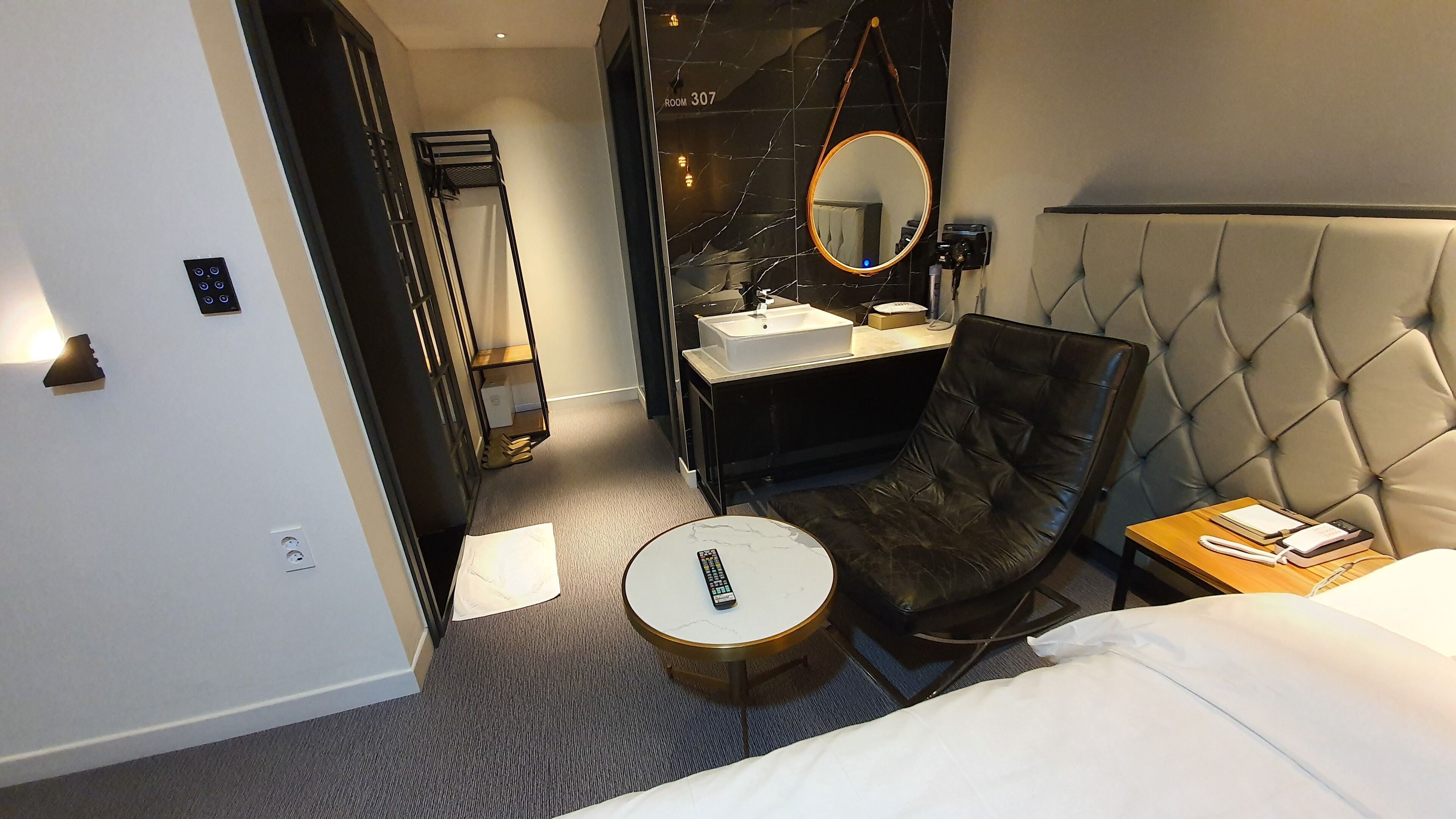 A hotel room with a bed, a round coffee table, a leather armchair, a bedside table with a phone, a mirror, a vanity with a sink, and a black-framed wardrobe.