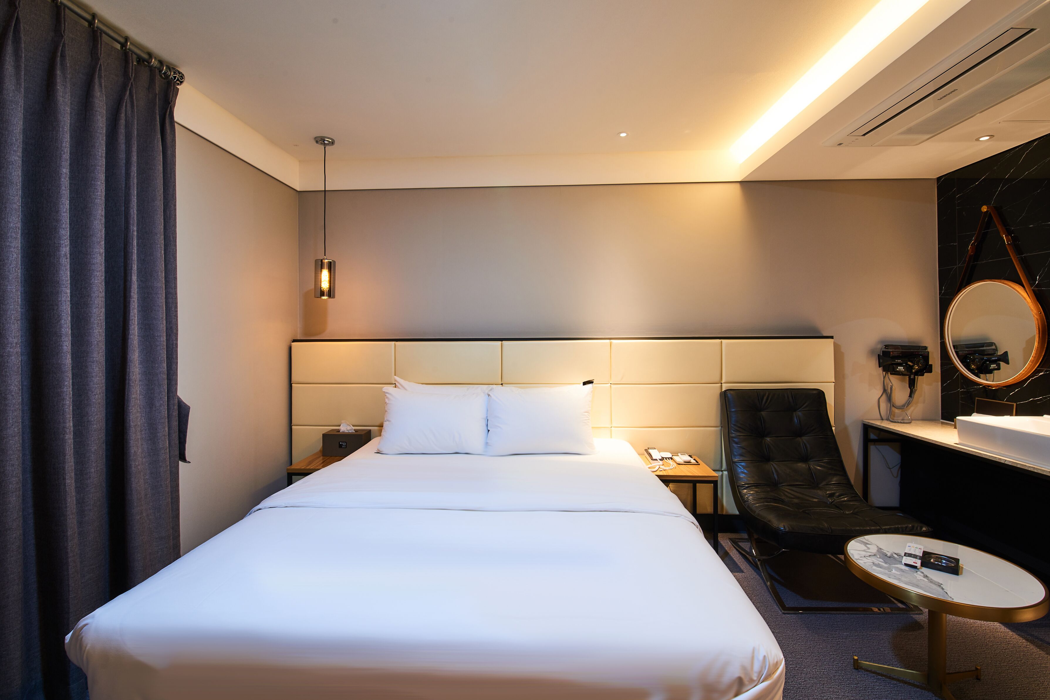 A hotel room with a large bed, a bedside table, a chair, a round table, a mirror, and a bathroom with a sink and mirror.