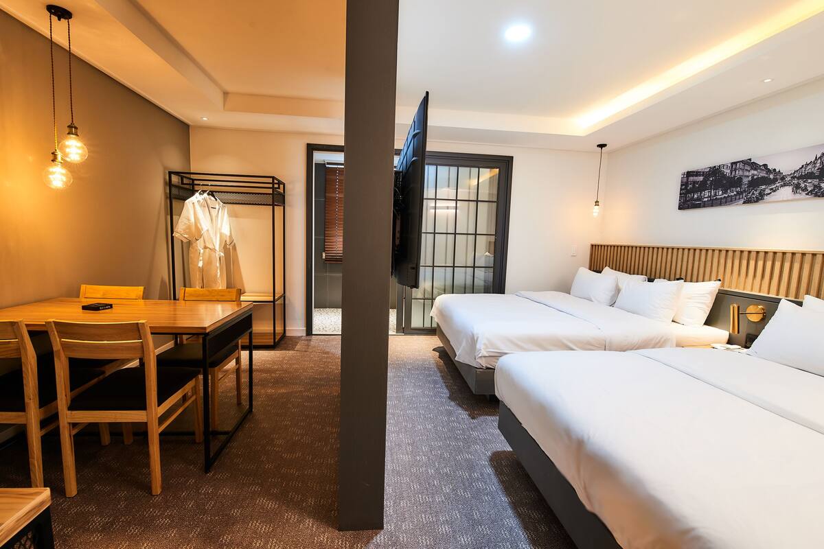 Una habitación de hotel con una mesa de comedor de madera, dos camas con ropa blanca, un televisor y un cuadro en la pared.