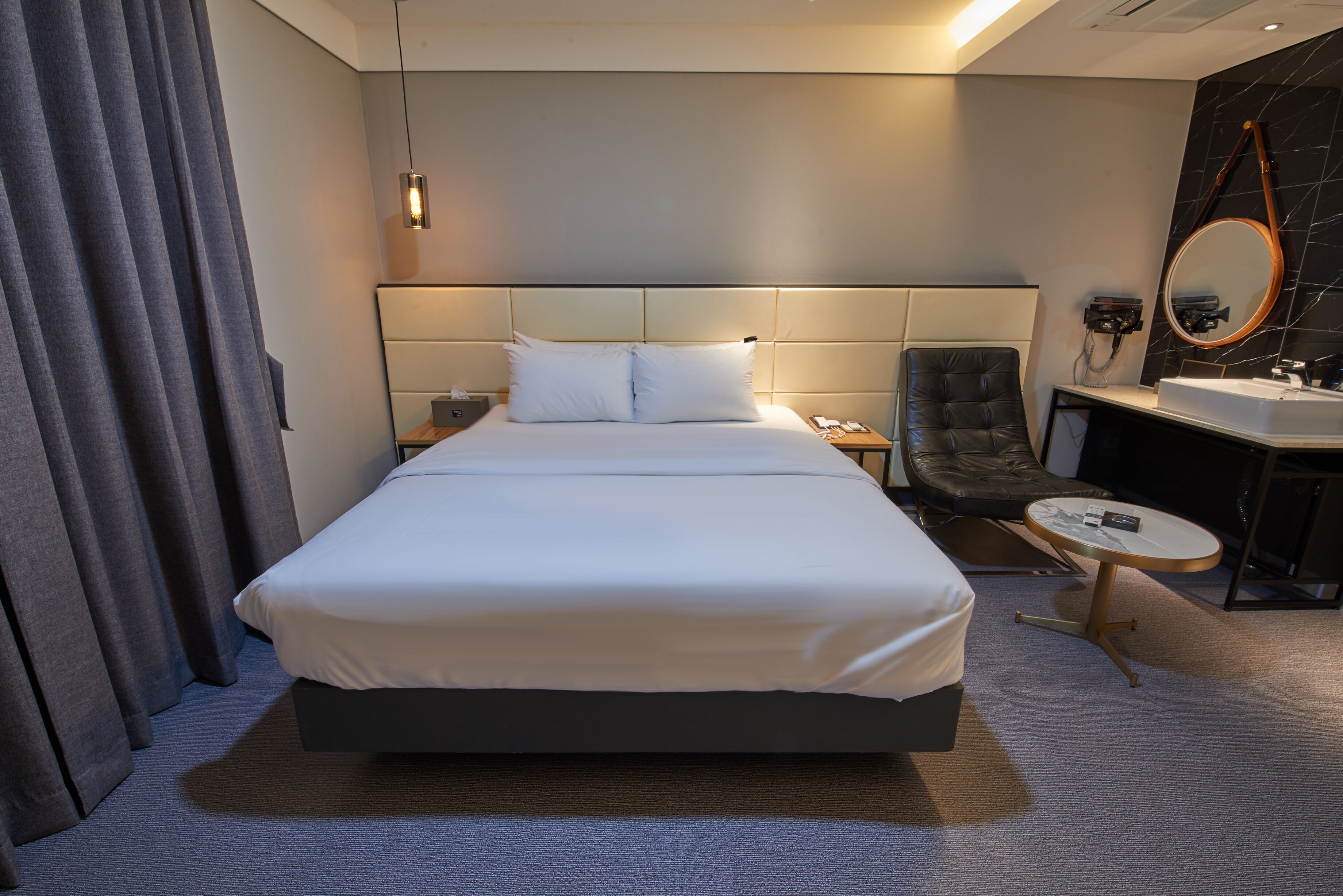 Una habitación de hotel con una cama grande, una mesita de noche, una silla, una mesa redonda, un espejo y un baño con lavabo y espejo.