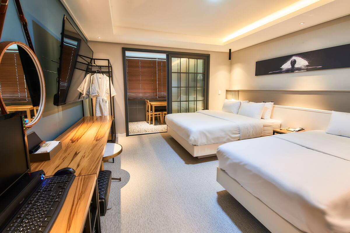 Una habitación de hotel moderna con un escritorio de madera, dos camas, un espejo grande y un ventanal corredero que da a un balcón.