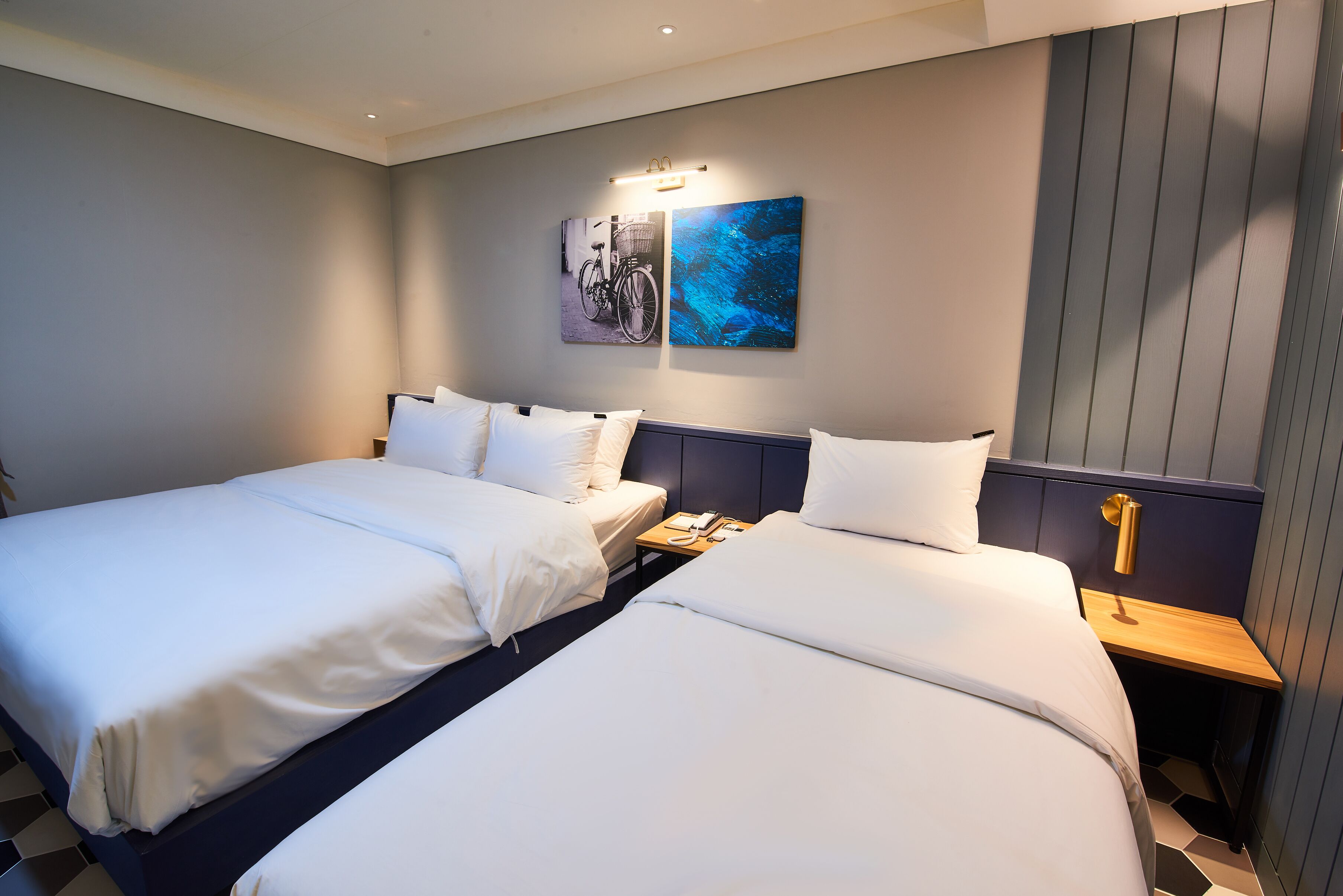 A hotel room with two beds, a nightstand, a wall-mounted lamp, and two framed pictures.