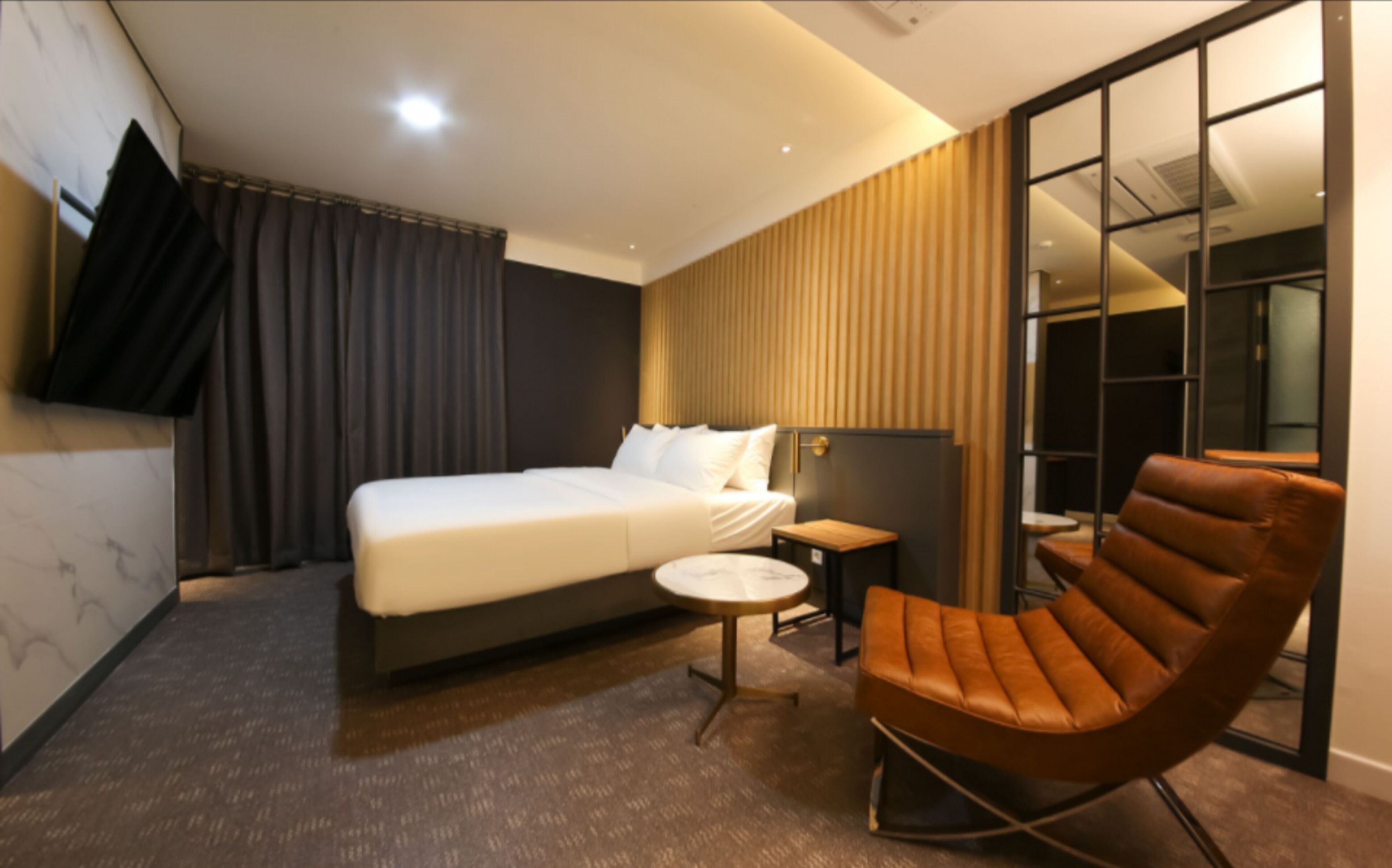 A modern hotel room with a large bed, a small round table, a leather armchair, and a large window with blinds.