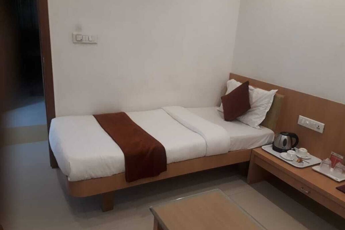A single bed with a brown blanket, a bedside table with a kettle, cups, and a tray, and a small table in front of the bed.