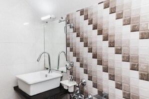 A bathroom with a white sink, two faucets, a mirror, and a tiled shower wall. - Hotel Amber (Vadodara)