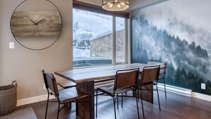 A dining area with a wooden table and chairs, a large wall clock, and a view of a snowy landscape through a window. - Cozy Ski-In/Ski-Out Escape: 3BR Echo Landing Home Near Orient Chairlift Sleeps 7 (Sun Peaks)
