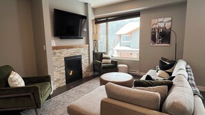 A modern living room with a fireplace, a large flat-screen TV, a sofa, and a window with a view of a house. - Cozy Ski-In/Ski-Out Escape: 3BR Echo Landing Home Near Orient Chairlift Sleeps 7 (Sun Peaks)