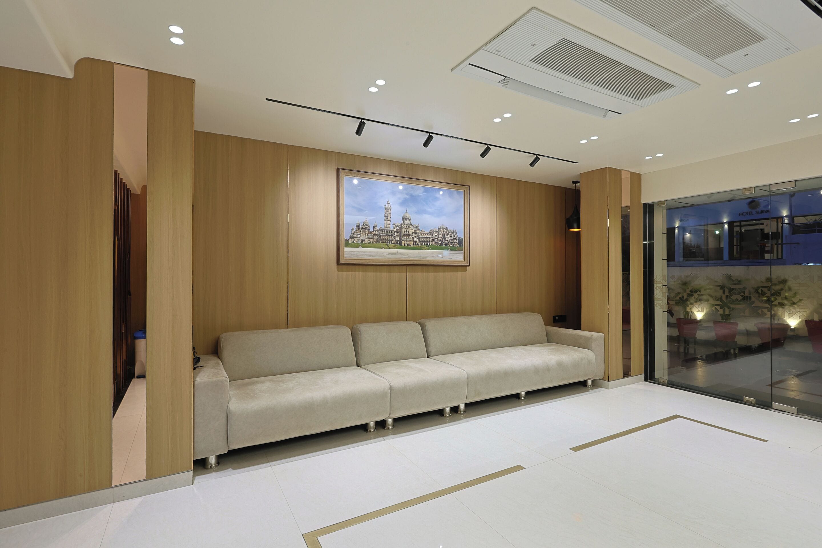 A modern hotel lobby with a beige sofa, a framed cityscape painting, and a glass entrance to the dining area.