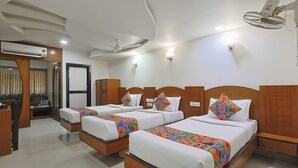 Habitación de hotel con tres camas, cada una con una colcha de diseño geométrico y colores vivos, cabeceros de madera y ropa de cama blanca. Se observa un ventilador de techo, un equipo de aire acondicionado y un escritorio pequeño con una silla.