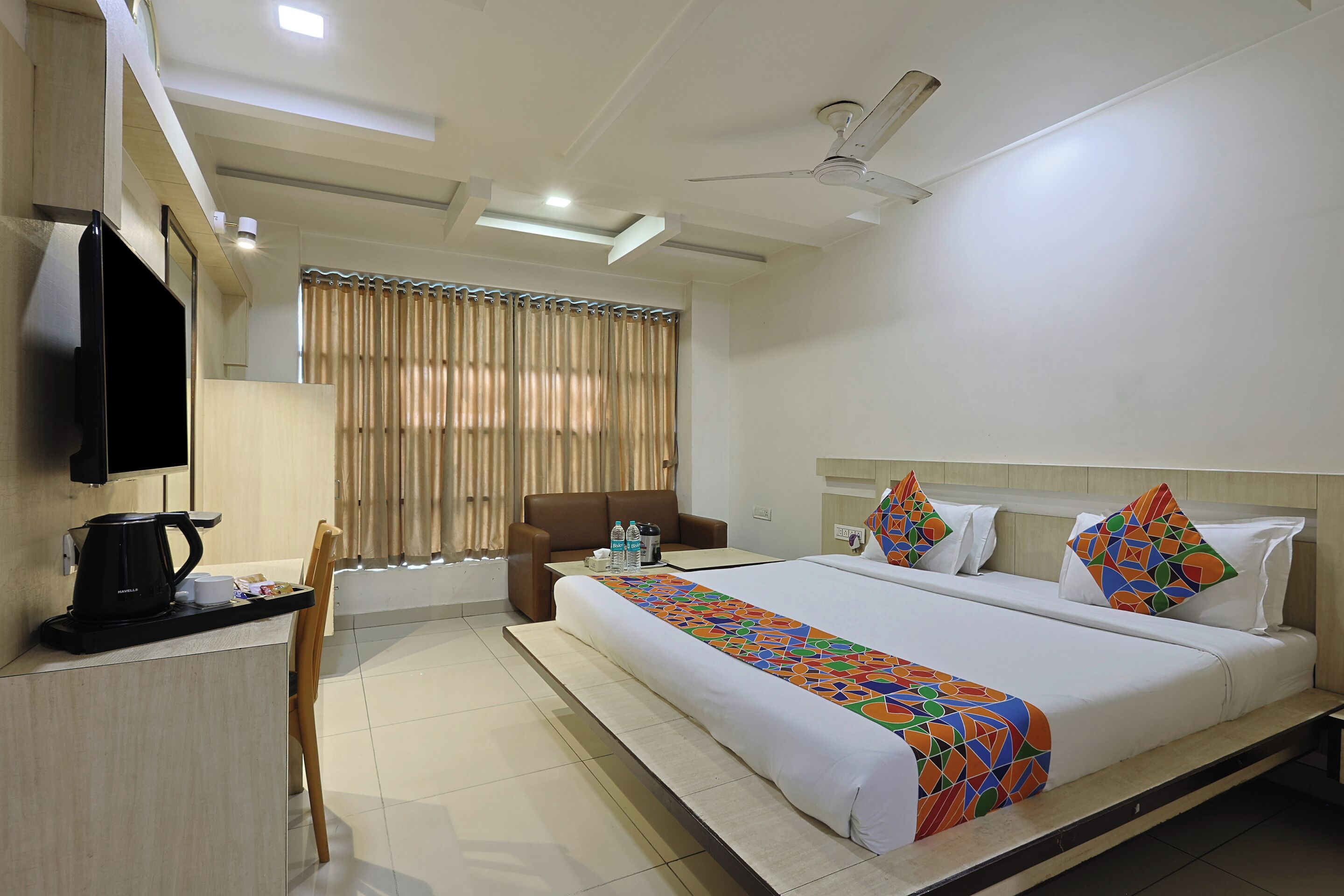 A hotel room with a bed, a desk with a coffee maker, a chair, a TV, and a window with curtains.