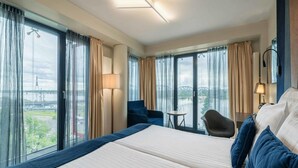 A hotel room with a large bed, a chair, a small table, and a view of a bridge and greenery. - Wellton Riverside SPA Hotel (Riga)
