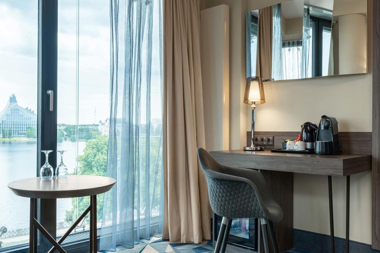 A hotel room with a large window, a round table with two glasses, a chair, a desk with a lamp, a coffee maker, and a mirror.