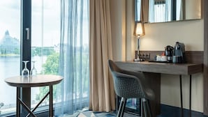 A hotel room with a large window, a round table with two glasses, a chair, a desk with a lamp, a coffee maker, and a mirror. - Wellton Riverside SPA Hotel (Riga)