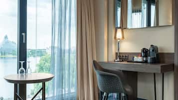 A hotel room with a large window, a round table with two glasses, a chair, a desk with a lamp, a coffee maker, and a mirror.