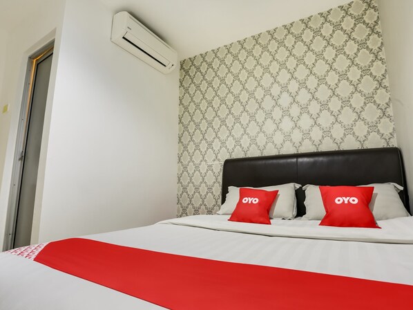 A hotel room with a bed, two red pillows with 'OYO' branding, a patterned wallpaper, and an air conditioning unit on the wall.