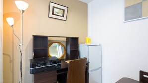 A compact kitchenette with a microwave, stove, and refrigerator. There is a wooden chair and a small table. A framed picture hangs on the wall above the mirror. - Sakamoto Building 7F (Osaka)