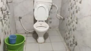 Ein kleines Badezimmer mit Fliesen, einer Toilette, einem grünen Eimer und einem blauen Hocker.
