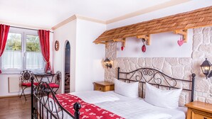 Een slaapkamer met een stenen muur, een houten plafond, een bed met wit beddengoed, een rode en witte gestreepte plaid, een nachtkastje, een houten stoel, een tafel en een raam met rode gordijnen.
