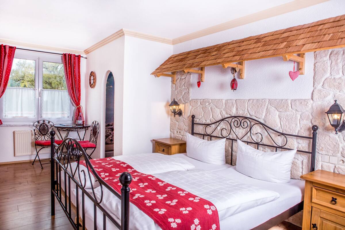 Ein Schlafzimmer mit Steinsandsteinwand, Holzbalkendecke, einem Bett mit weißer Bettwäsche, einer rot-weißen Decke mit Muster, einem Nachttisch, einem Holzstuhl, einem Tisch und einem Fenster mit roten Vorhängen.