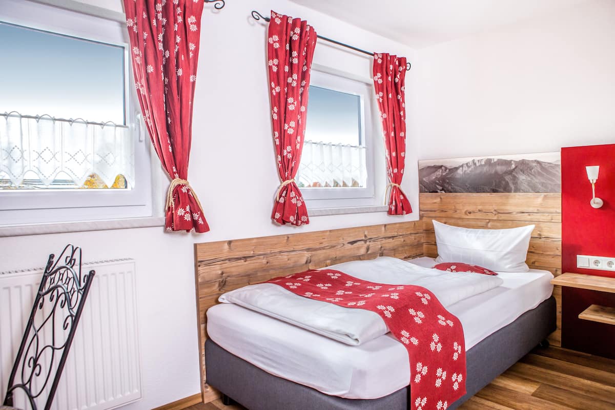 Ein Schlafzimmer mit einem Bett, einem Holz-Kopfbrett, roten Vorhängen, einem Fenster mit Aussicht und einer roten Wand mit Lichtschalter.