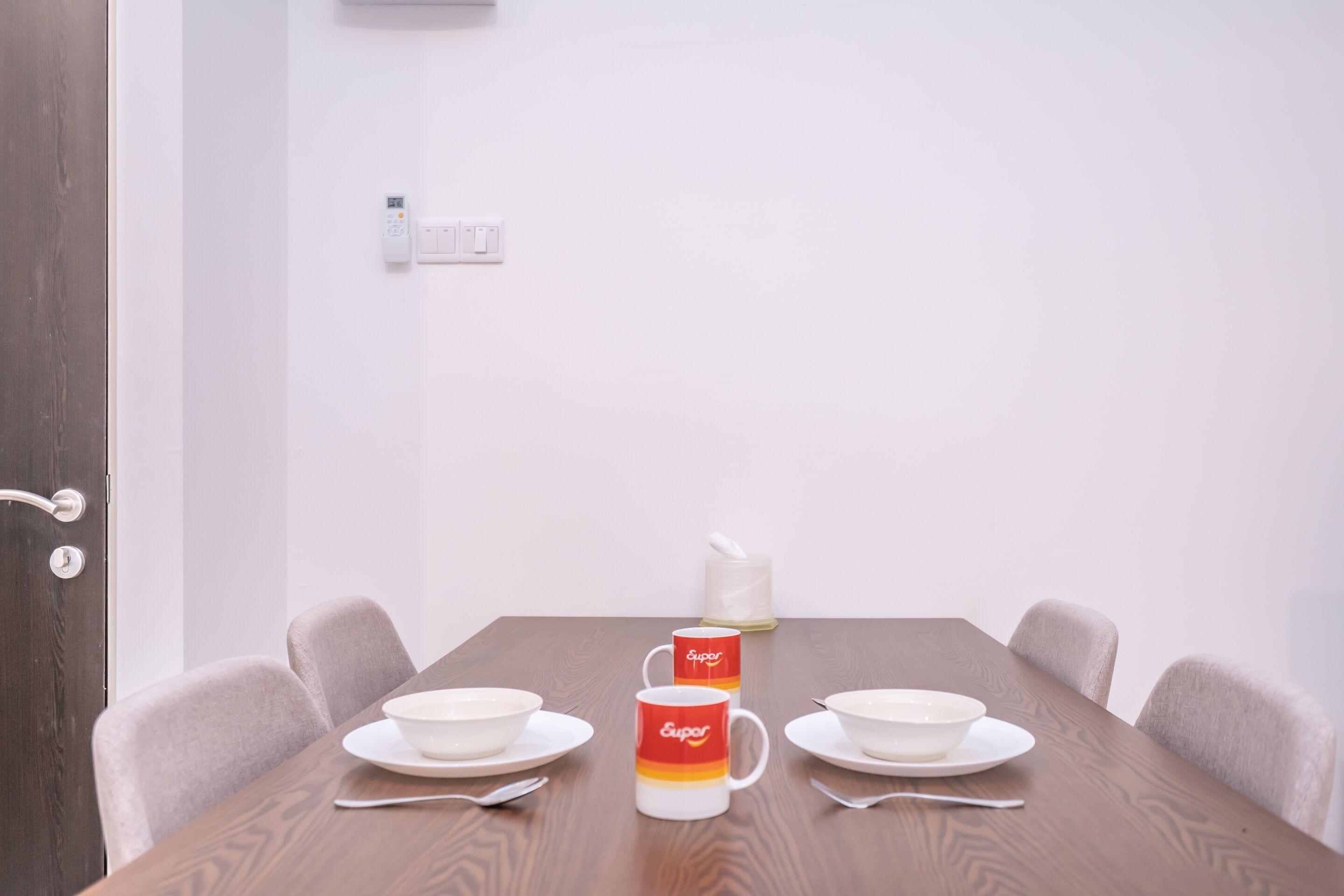 Een eettafel met twee witte kommen, twee witte borden en twee lepels. Er staat een rode en gele mok met de tekst 'Eupor' erop. De tafel staat voor een witte muur met een lichtschakelaar erboven.