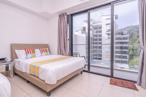 Una habitación de hotel moderna con una cama grande, una mesita de noche, un ventanal con vistas a edificios y vegetación, y una puerta corrediza de vidrio.
