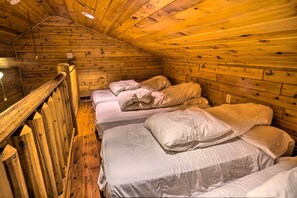 Una cabaña con paredes de madera y techo inclinado. Se observan tres camas con ropa blanca.