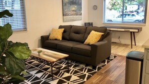 Un salon avec un canapé marron, deux coussins à motifs, une table basse, une grande plante, un tableau accroché au mur et une fenêtre donnant sur la rue.
