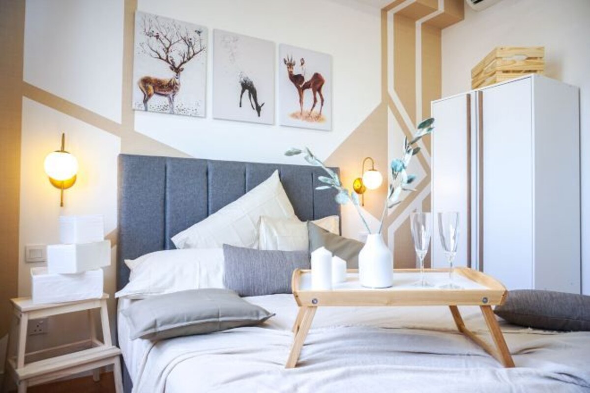 A modern bedroom with a grey upholstered headboard, a bedside table with a lamp, a tray with glasses and candles, and wall art depicting deer.
