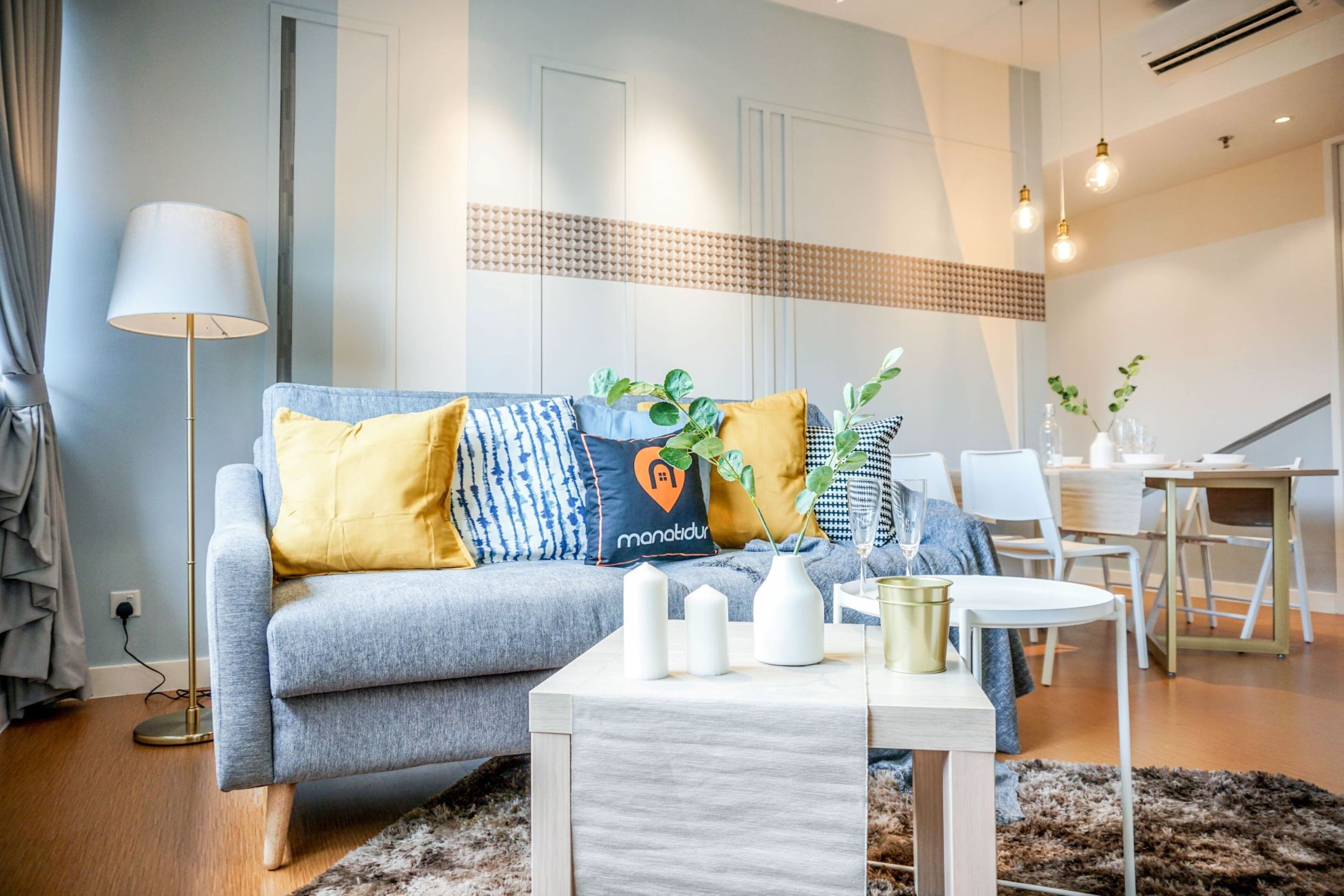 A modern living room with a grey sofa, yellow and patterned cushions, a small white table with candles, and a dining area with a white table and chairs.