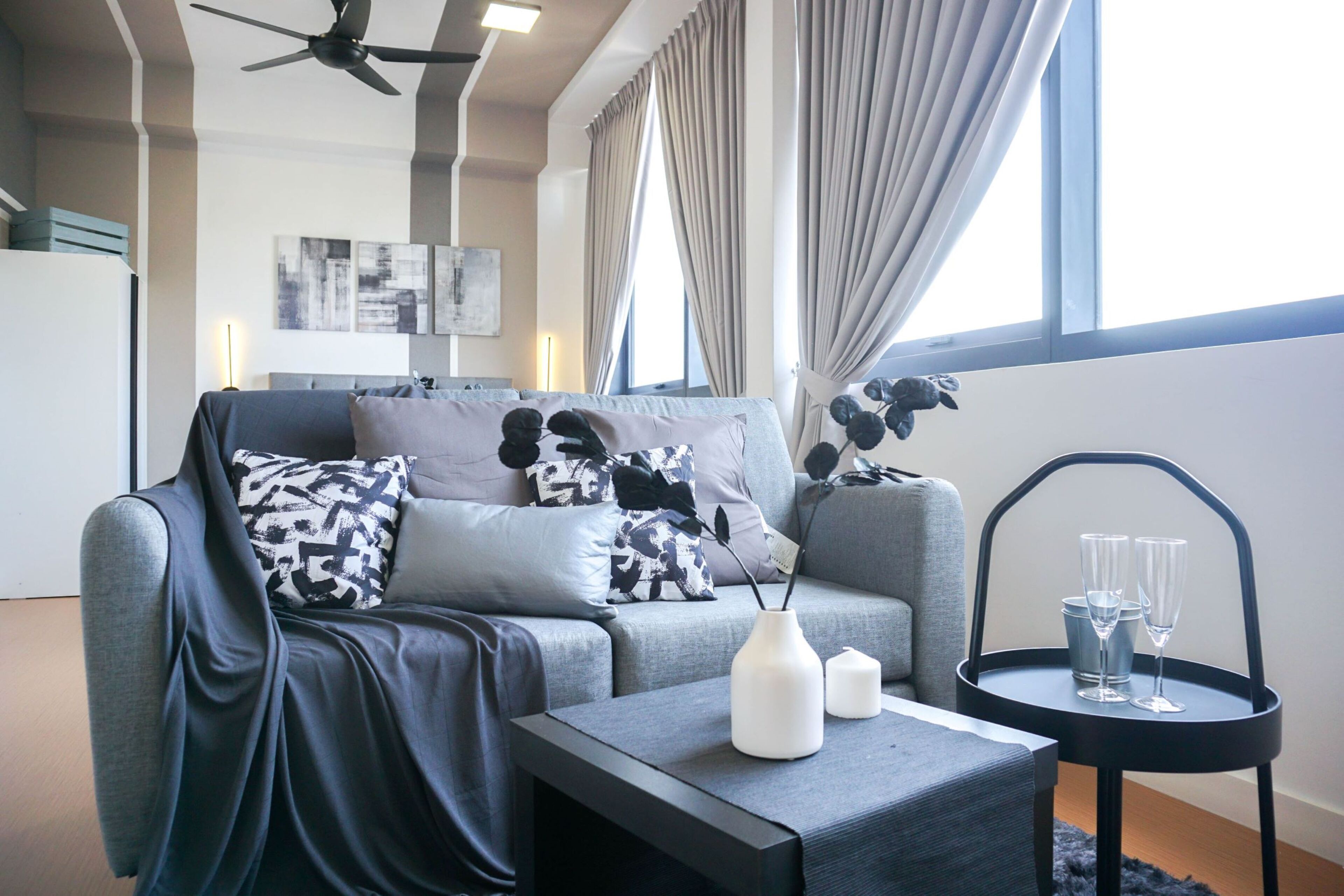 Una sala moderna con un sofá gris, almohadones estampados en blanco y negro, una mesa de centro negra con un jarrón blanco y velas, y una mesita con dos vasos.