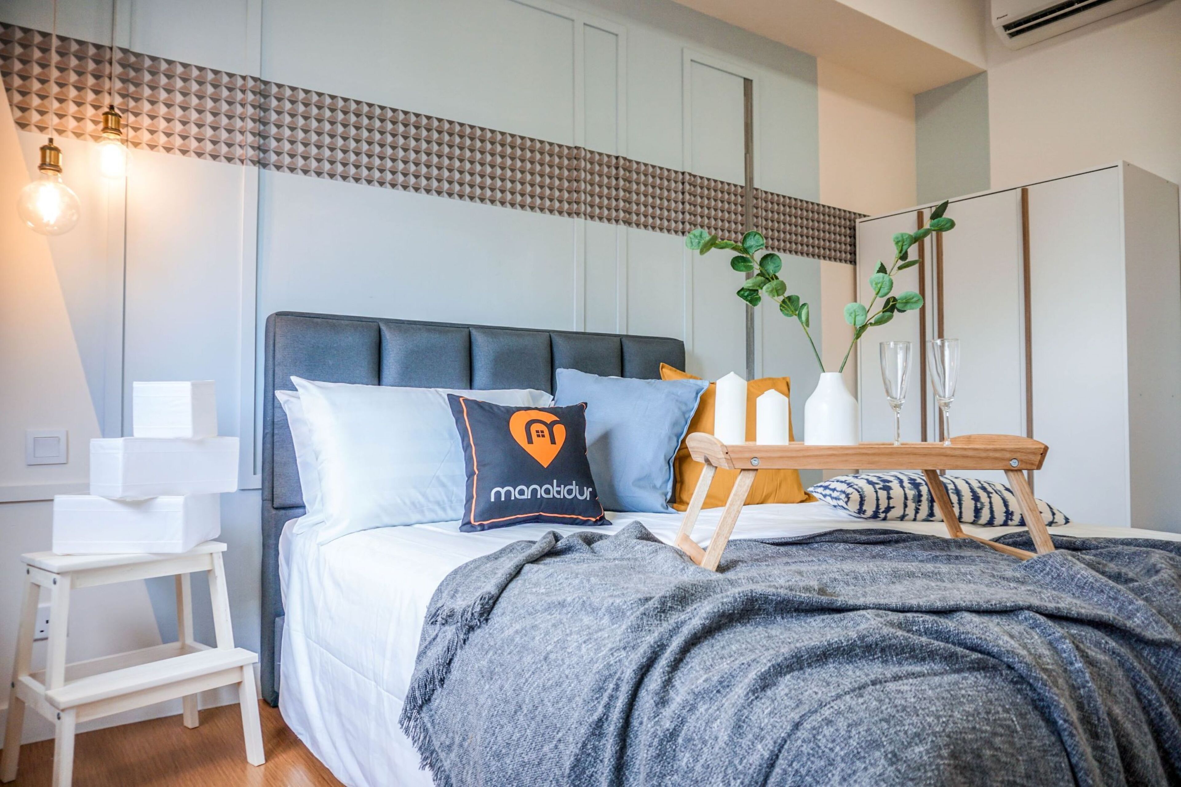 A modern bedroom with a grey upholstered bed, a bedside table with a lamp, and a mirror on the wall.
