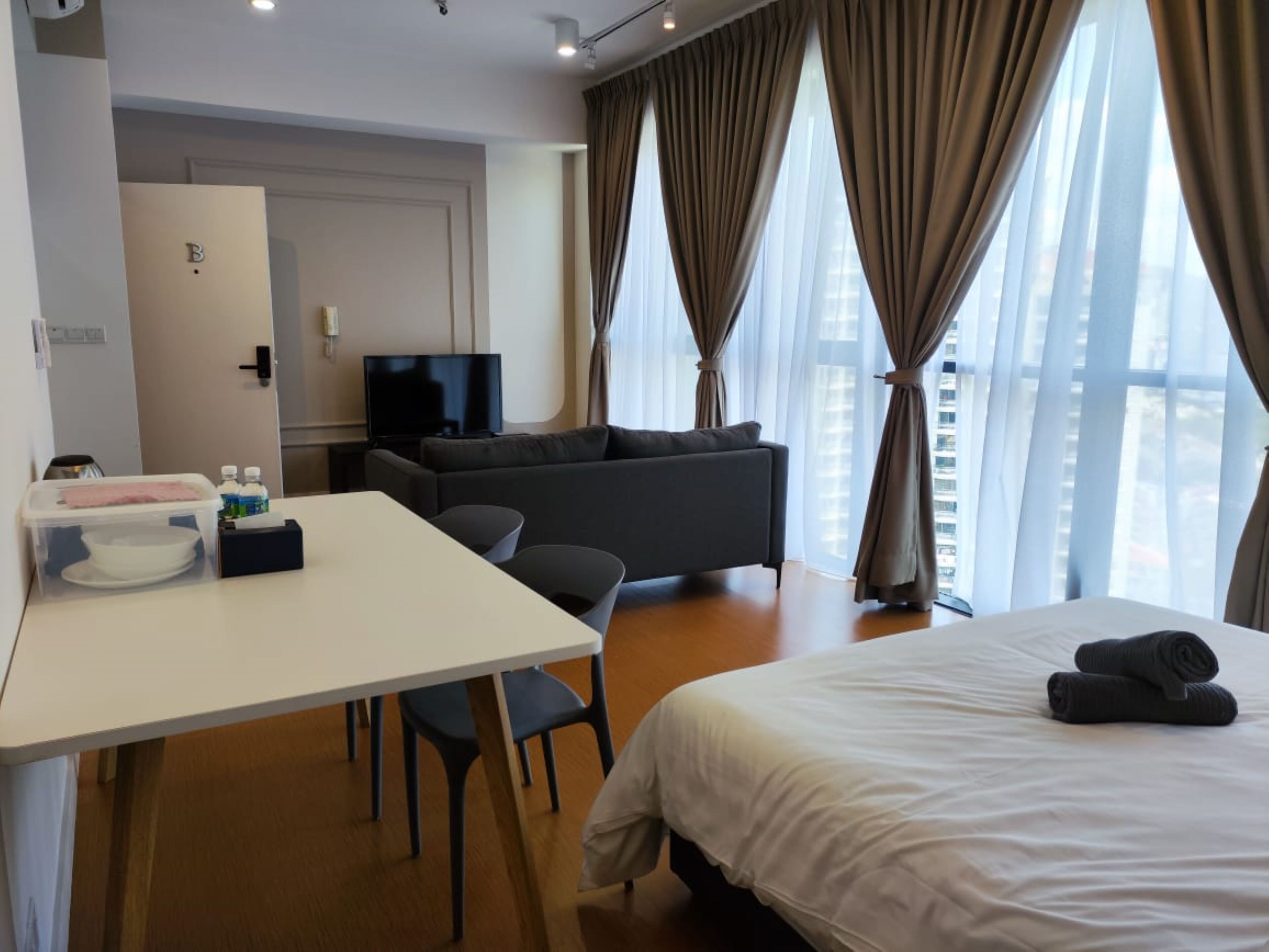A hotel room with a bed, a sofa, a TV, a desk with chairs, a mini-fridge, and a window with curtains.