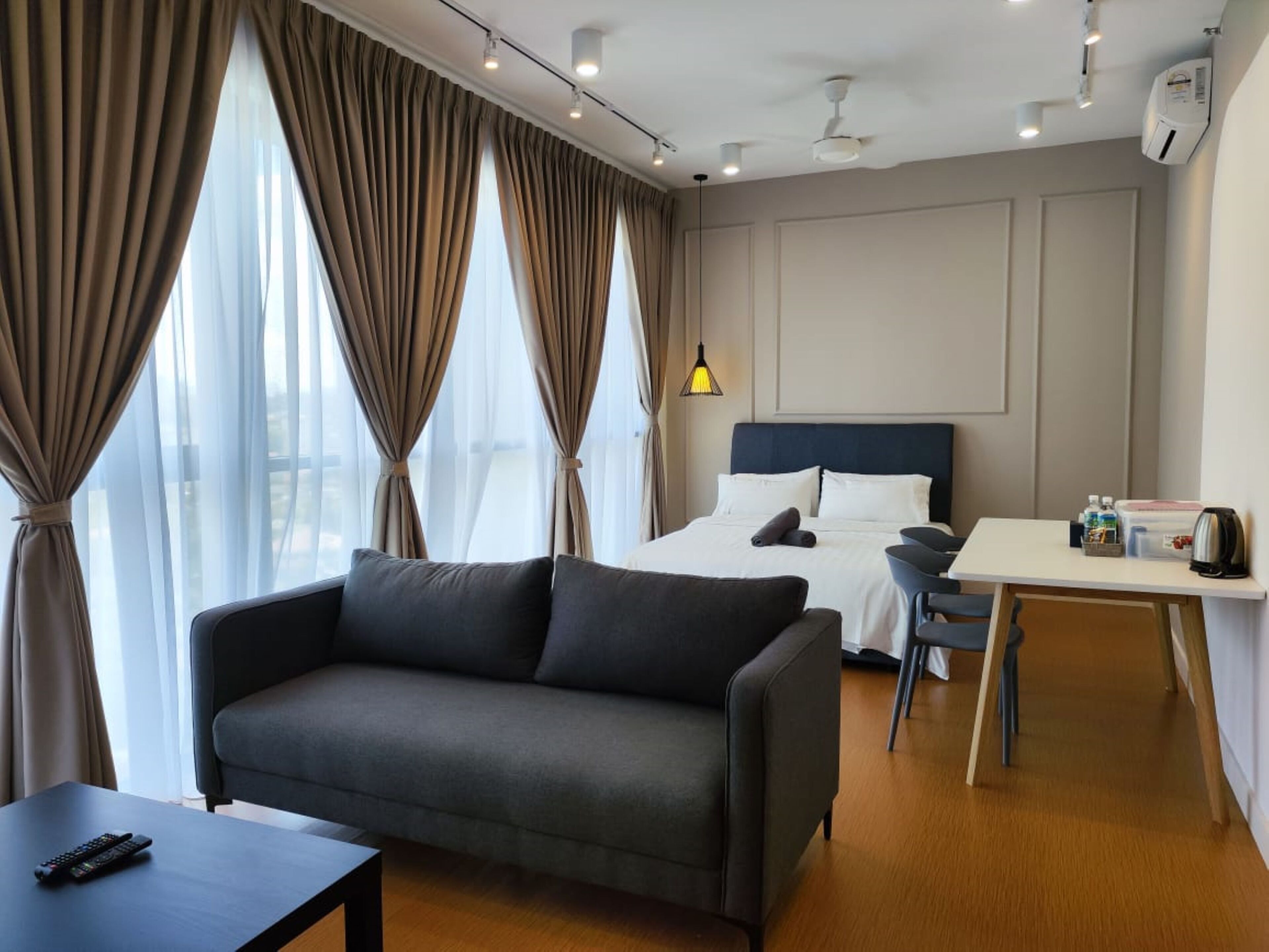 A hotel room with a bed, a sofa, a desk, and a chair. There is a window with curtains, a ceiling fan, and a wall-mounted air conditioner.
