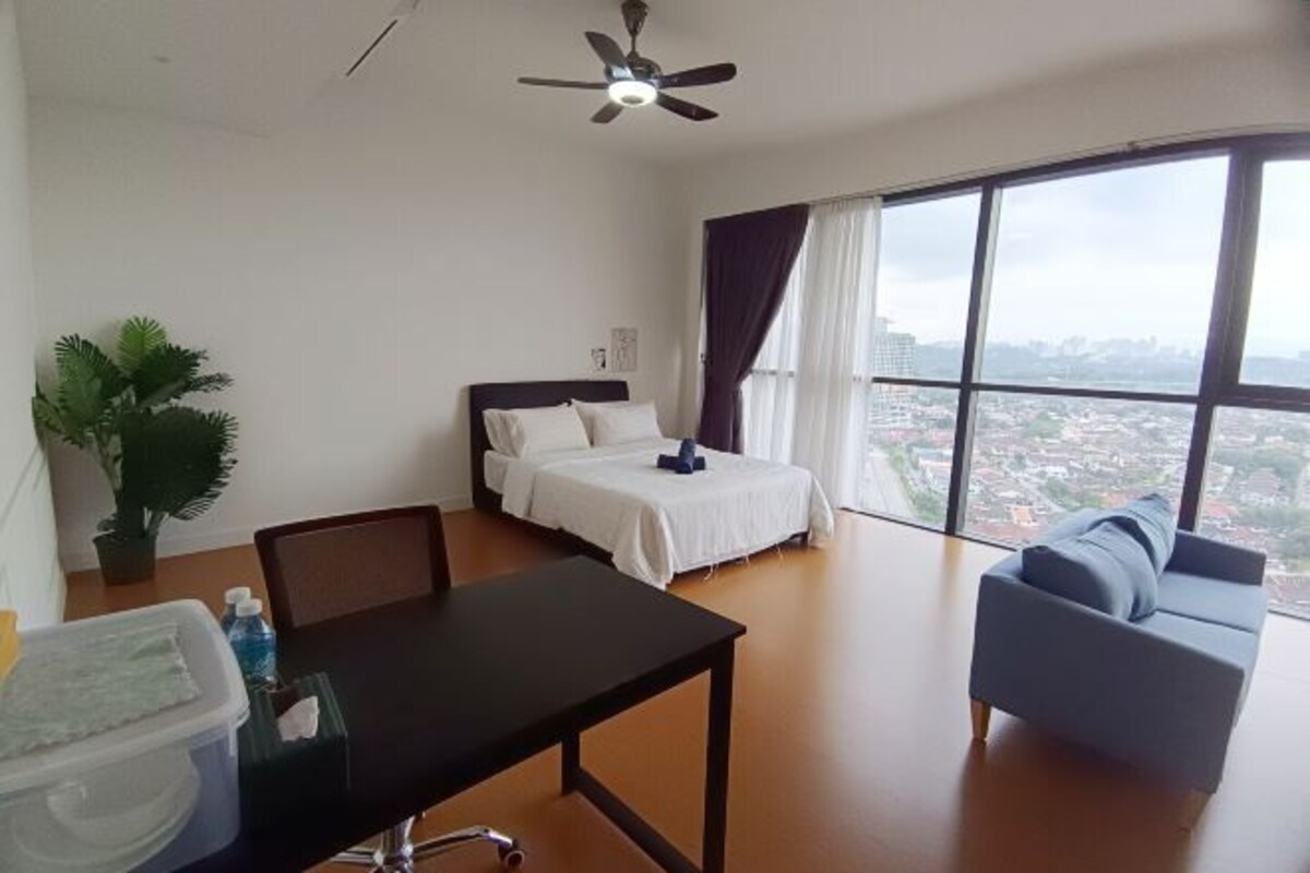 A modern hotel room with a bed, a desk, a chair, a sofa, a ceiling fan, a large window with a city view, and a potted plant.