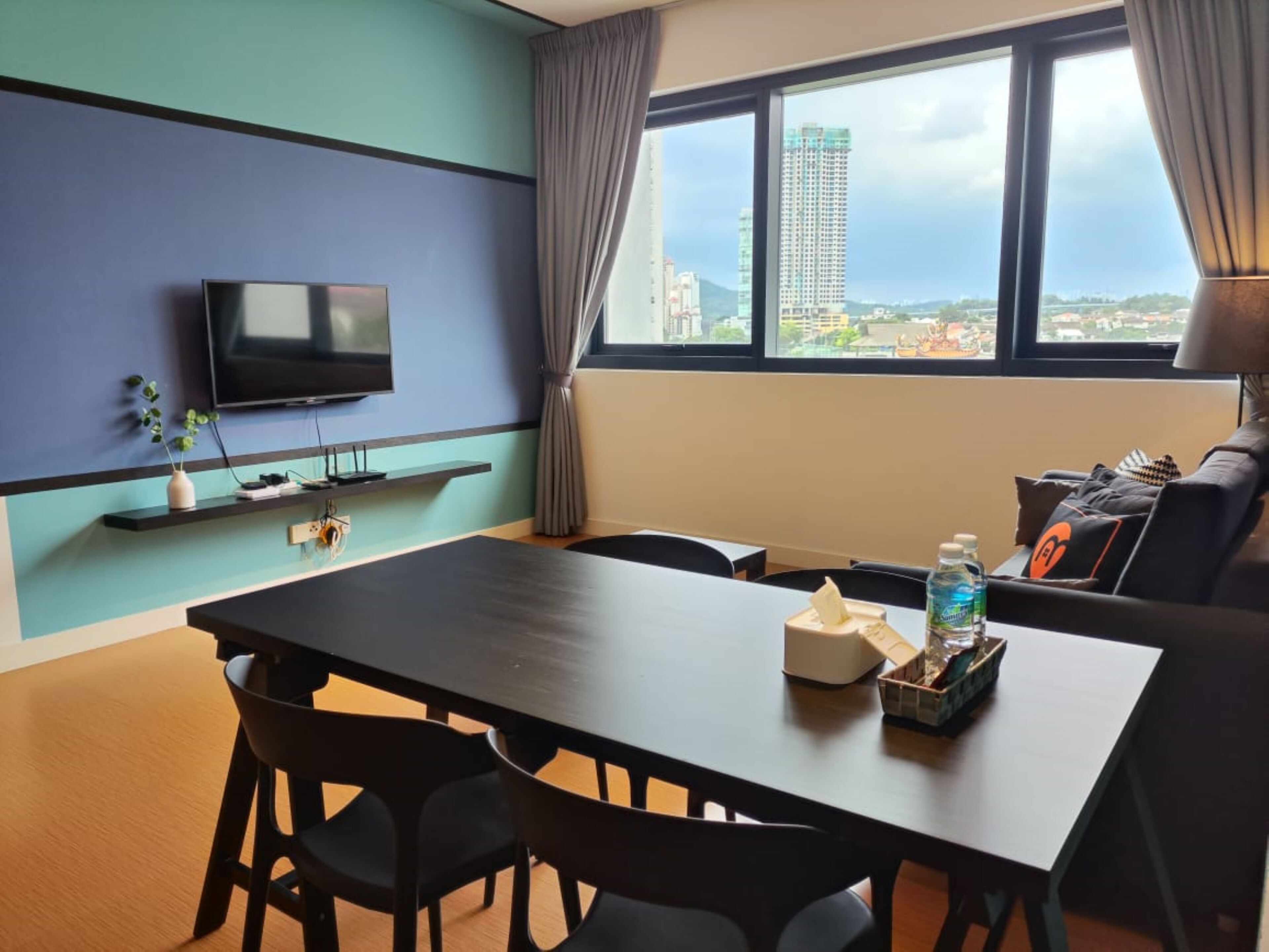 A modern hotel room with a dining area, a flat-screen TV mounted on the wall, a window with a city view, and a sofa with a table beside it.