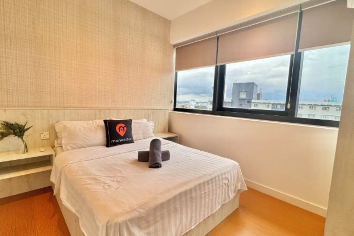 A hotel room with a large bed, a nightstand, a window with blinds, and a view of buildings outside.