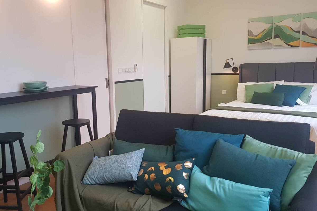 Un espace de vie compact comprenant un coin cuisine, un lit avec des oreillers verts et bleus, une plante et un tableau accroché au mur.