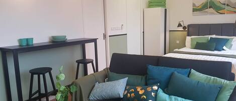 A compact living space with a kitchenette, a bed with green and blue pillows, a plant, and a painting on the wall.