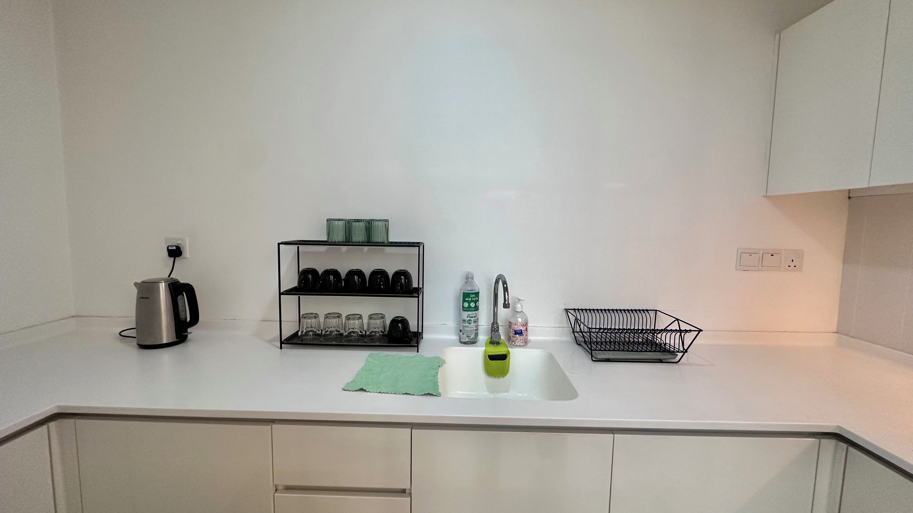 Una cocina con encimera blanca, hervidor, estante con vasos, fregadero y grifo.