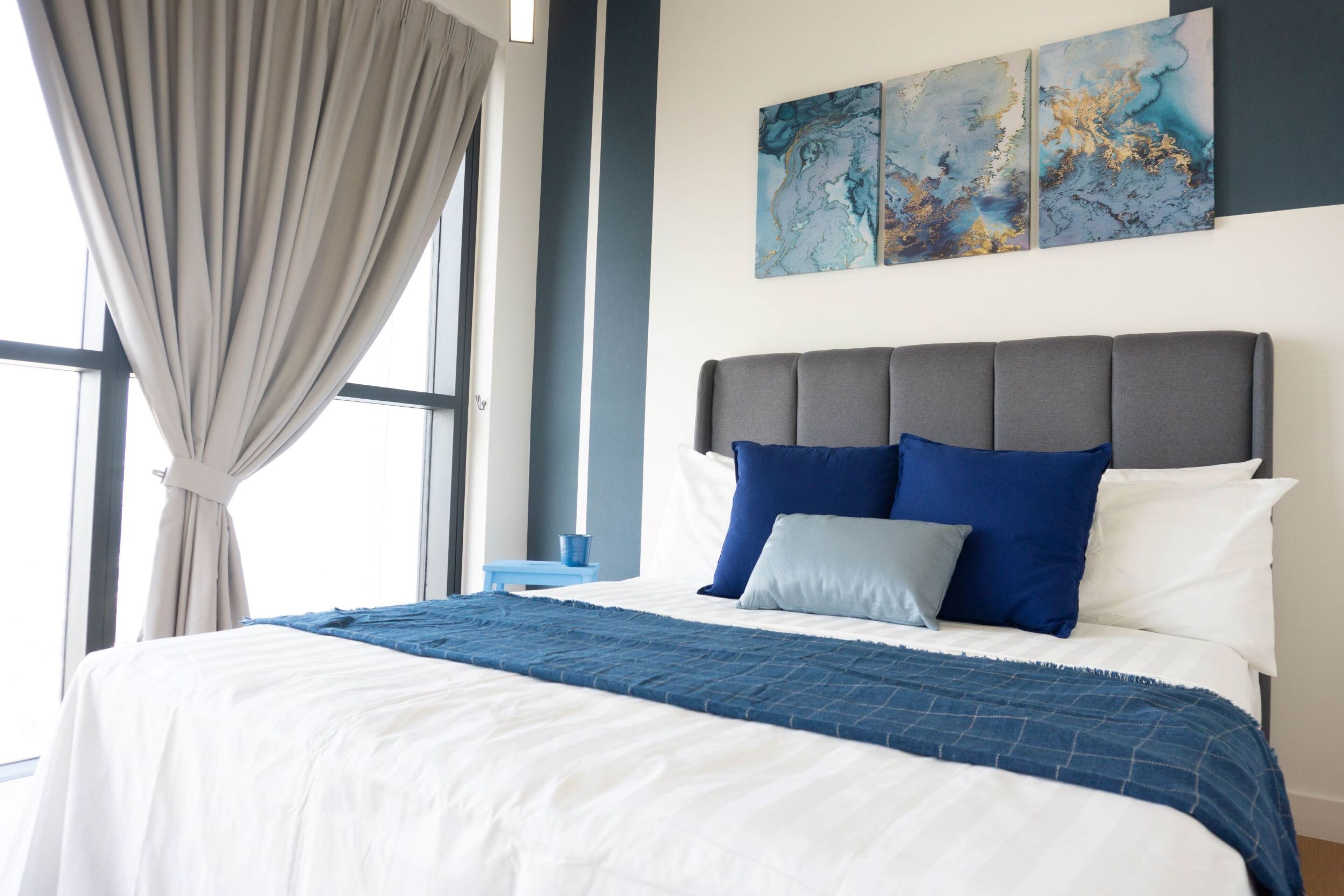 A neatly made bed with a blue and white bedspread, a grey upholstered headboard, and two blue pillows.