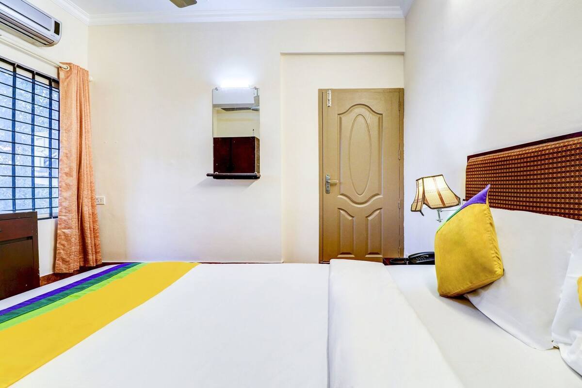 A hotel room with a bed, a door, a wall-mounted lamp, and a window with blinds.