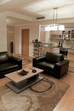 Un salon moderne avec deux canapés en cuir noir, une table basse en bois et un grand tapis. On aperçoit, au fond, une cuisine équipée d’appareils électroménagers en acier inoxydable.